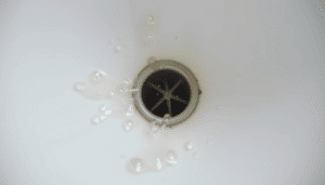 Close-up of a clogged bathroom sink drain with pooled water, soap scum, and hair on white porcelain in natural light.